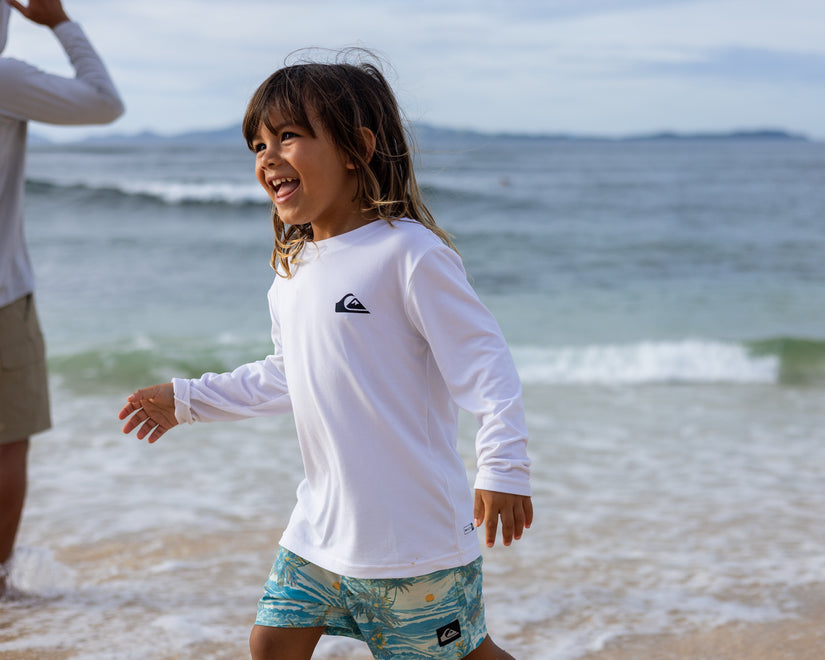 Child wearing a white long-sleeve shirt with a logo and colorful shorts on a beach.