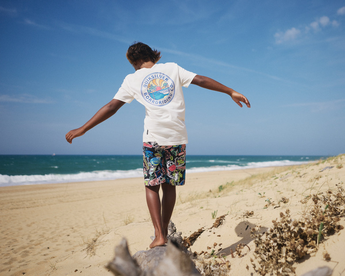 Person standing on a beach with arms outstretched, wearing a white t-shirt and colorful shorts.