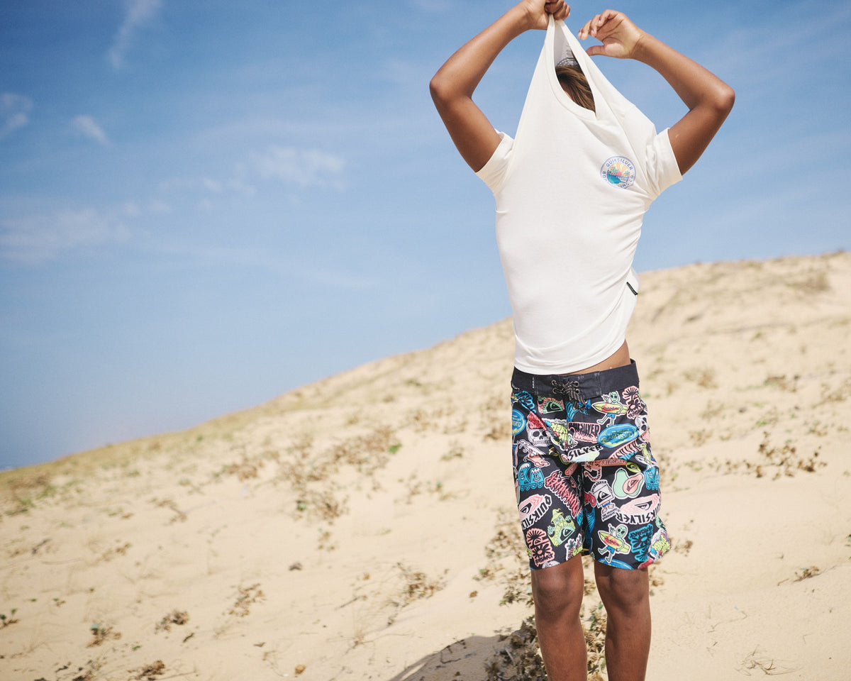 Person wearing a white t-shirt and colorful shorts on a sandy beach with blue sky.