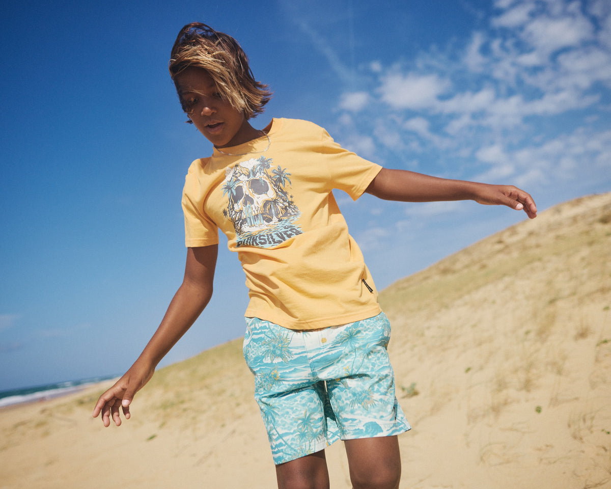 Person wearing a yellow t-shirt and blue shorts standing on a sandy beach with a clear blue sky.