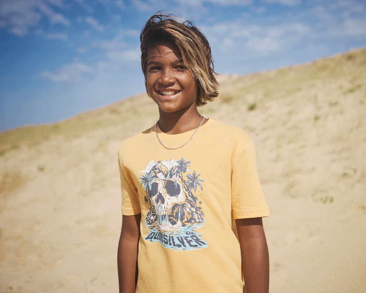 Child wearing a yellow t-shirt with a graphic design, standing on sand dunes under a blue sky.