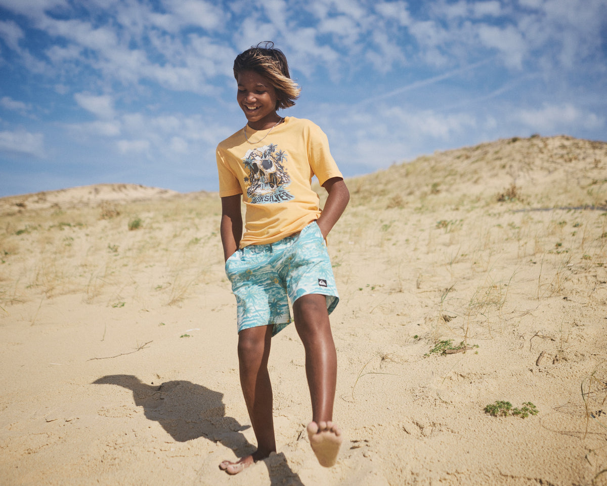 Child in yellow t-shirt and blue shorts standing on a sandy dune with a blue sky.